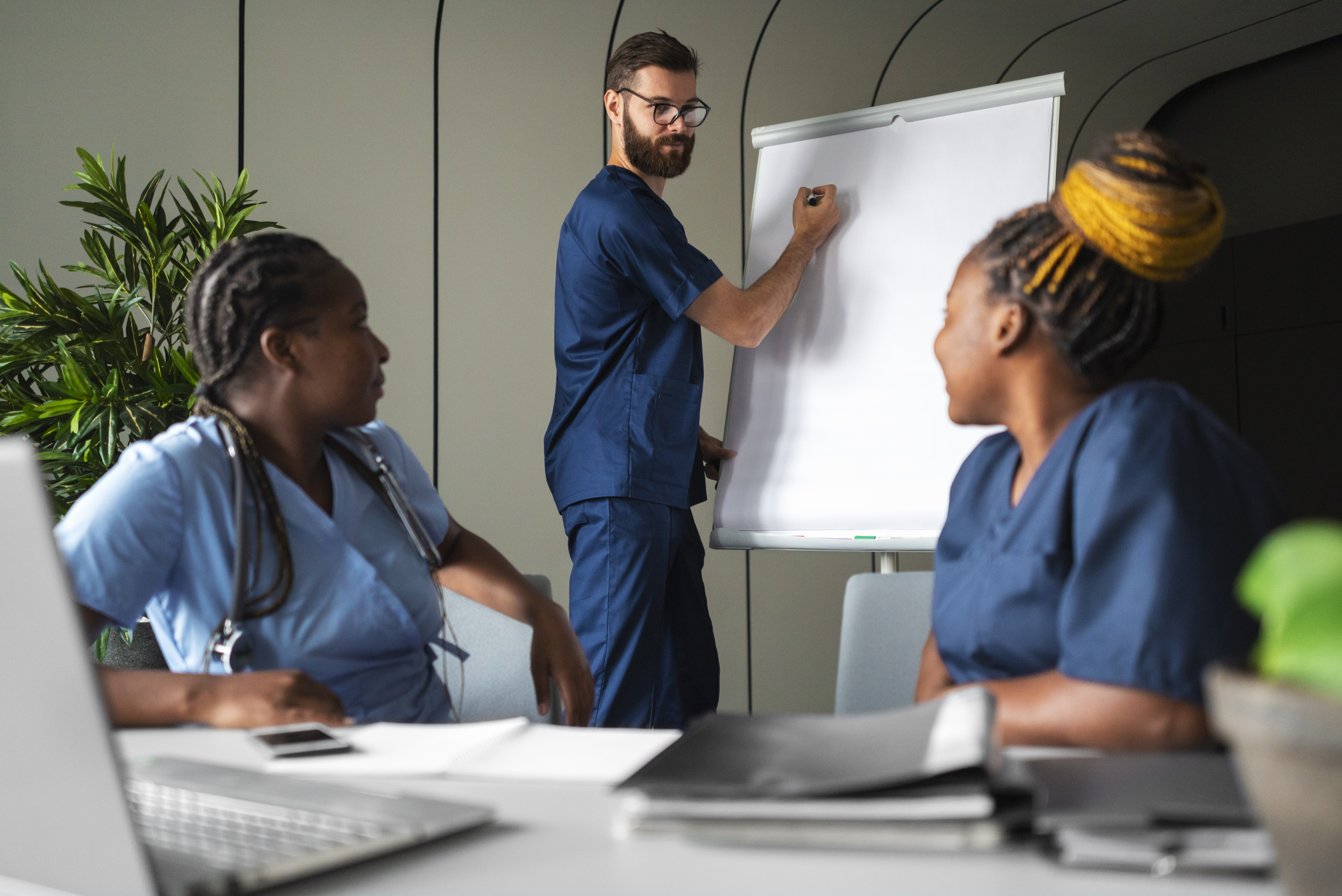 Side view of three healthcare professionals in blue scrubs conducting a workshop; a bearded male nurse stands writing on a whiteboard flip chart while two female nurses sit at a desk with notes and a laptop listening attentively.