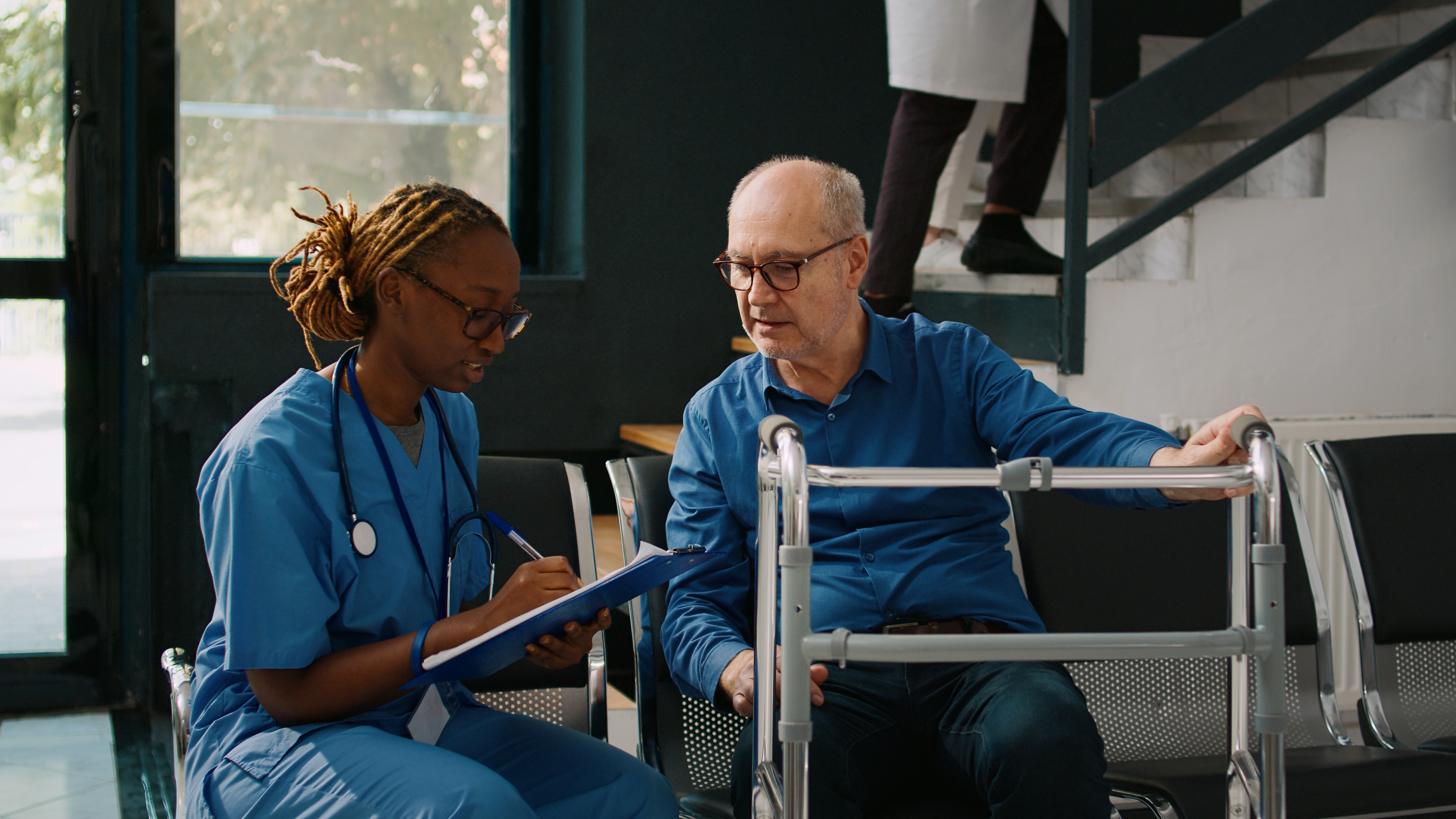 A female nurse in blue scrubs writing on a clipboard while seated next to a senior man holding a walking frame.